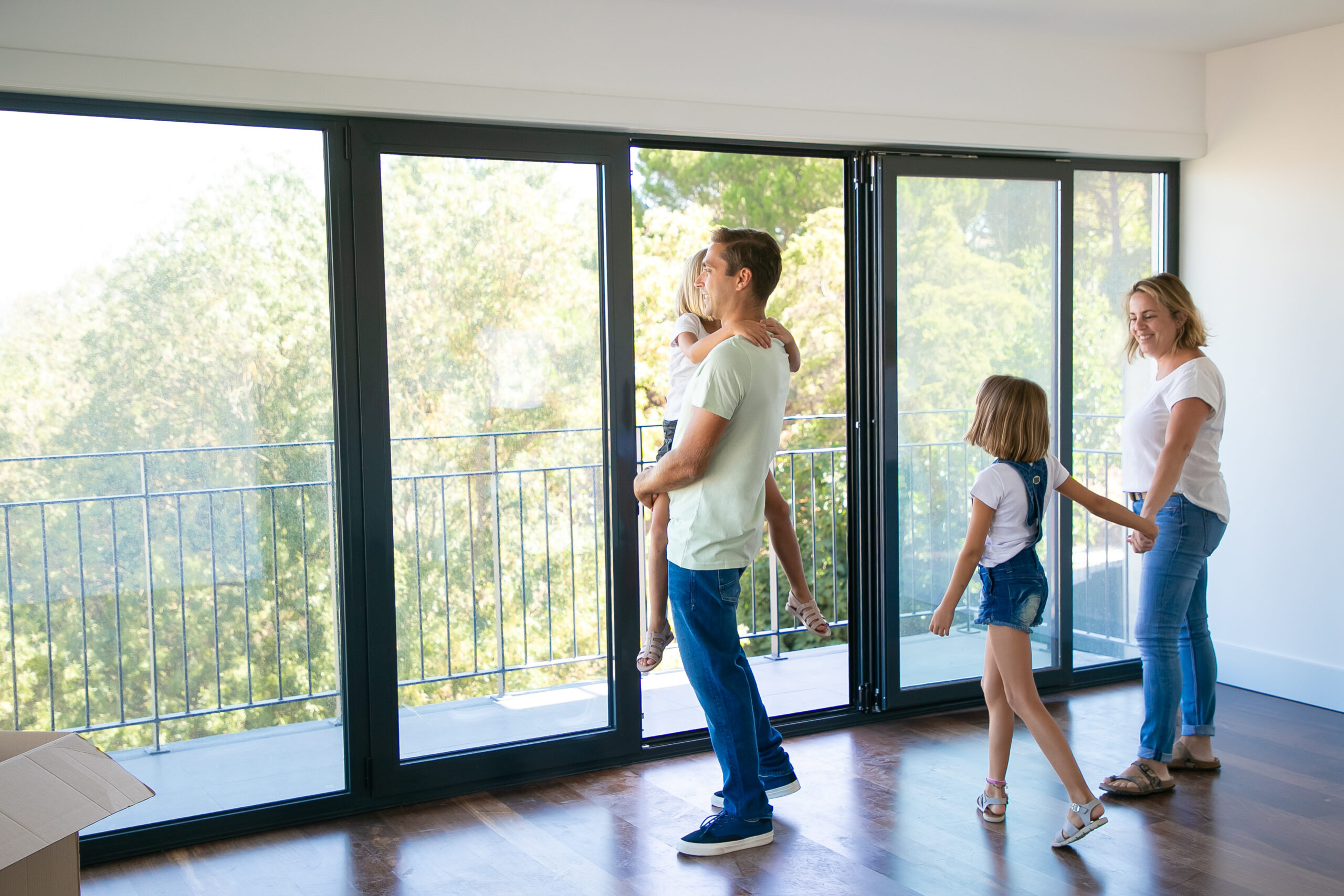 Happy father with daughter standing near open balcony and smiling. Blonde mother holding girl hand and talking with her. Enjoying family watching new house or flat. Relocation and mortgage concept